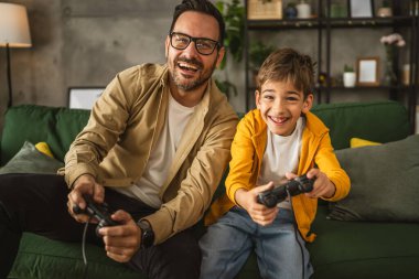 Baba ve oğlu beyaz tenli, joystick ile video oyunu oynuyor ve evde eğleniyor.
