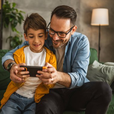 Baba ve oğul cep telefonuyla video oyunu oynuyor ve evde eğleniyorlar.