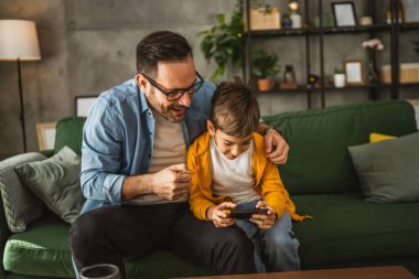 Baba ve oğul cep telefonuyla video oyunu oynuyor ve evde eğleniyorlar.