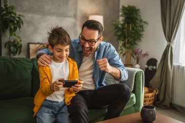 Baba ve oğul cep telefonuyla video oyunu oynuyor ve evde eğleniyorlar.
