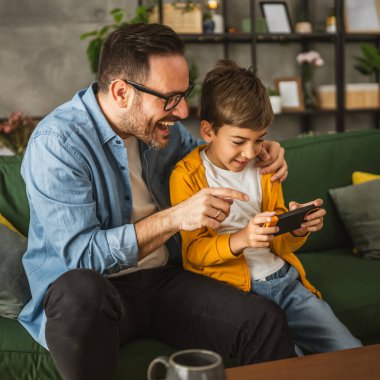 Baba ve oğul cep telefonuyla video oyunu oynuyor ve evde eğleniyorlar.