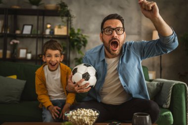 Baba ve oğlu Kafkas futbol maçı izleyip evde tezahürat yapıyorlar. Gürültülü, heyecanlı, mutlu.