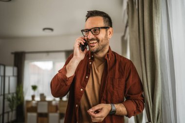 Yetişkin bir adamın portresi cep telefonuyla konuşurken ayakta durur ve evde mutlu bir şekilde gülümser.