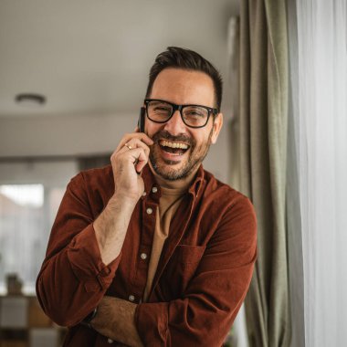 Yetişkin bir adamın portresi cep telefonuyla konuşurken ayakta durur ve evde mutlu bir şekilde gülümser.