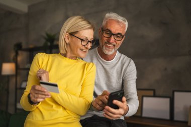 Olgun ya da yaşlı Kafkasyalı karı-koca internetten alışveriş yapıyor. Evde oturan koca cep telefonunu tutuyor ve kredi kartından bilgi okuyor.