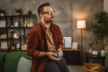 Yetişkin beyaz bir adam oturur ve elinde kahveyle evden dışarı bakar.