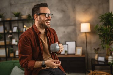 Yetişkin beyaz bir adam oturur ve elinde kahve ile pencereden dışarı bakar ve gülümser.