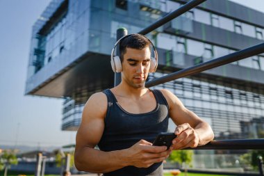 Beyaz tenli genç bir erkek açık hava eğitim parkı spor salonunda duruyor. Kulaklıklı cep telefonu kullanıyor. Gerçek insanları eğitmek için internetten mesajlar yolluyor.