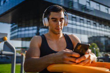 Beyaz tenli genç bir erkek açık hava eğitim parkı spor salonunda duruyor. Kulaklıklı cep telefonu kullanıyor. Gerçek insanları eğitmek için internetten mesajlar yolluyor.