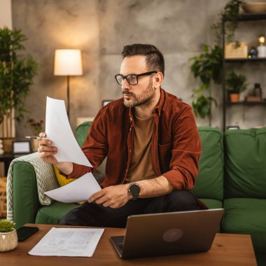 Gözlüklü yetişkin bir beyaz adam evde dizüstü bilgisayarla çalışıyor ve belgeleri tutuyor.