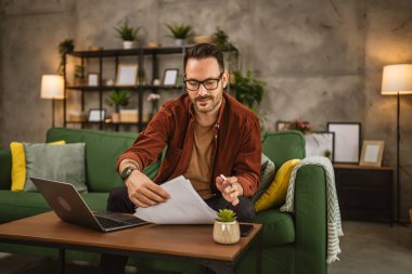 Gözlüklü yetişkin bir beyaz adam evde dizüstü bilgisayarla çalışıyor ve belgeleri tutuyor.