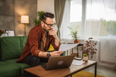 Gözlüklü yetişkin bir beyaz adam evde dizüstü bilgisayarla çalışıyor ve belgeleri tutuyor.