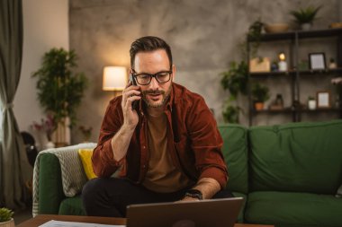 Gözlüklü yetişkin bir beyaz adam evde dizüstü bilgisayarla çalışıyor ve cep telefonuyla konuşuyor.