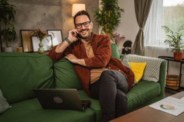 Gözlüklü yetişkin bir beyaz adam evde dizüstü bilgisayarla çalışıyor ve cep telefonuyla konuşuyor.