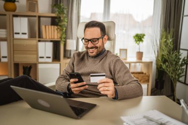 Erişkin işadamı işi bittikten sonra cep telefonuyla alışveriş yapıp kredi kartını tutuyor.