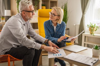 Olgun yaşlı çift karı koca birlikte çalışırlar. Evlerinde birlikte çalışırlar. Erkek ve kadın belgelere bakar ve konuşurlar.