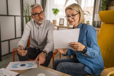 Olgun yaşlı çift karı koca birlikte çalışırlar. Evlerinde birlikte çalışırlar. Erkek ve kadın belgelere bakar ve konuşurlar.