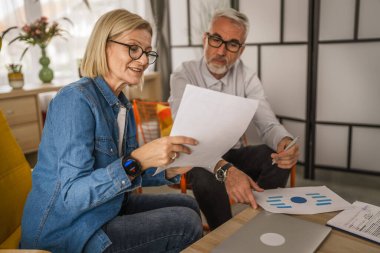 Olgun yaşlı çift karı koca birlikte çalışırlar. Evlerinde birlikte çalışırlar. Erkek ve kadın belgelere bakar ve konuşurlar.