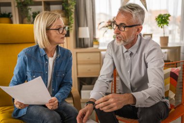 Olgun yaşlı çift karı koca birlikte çalışırlar. Evlerinde birlikte çalışırlar. Erkek ve kadın belgelere bakar ve konuşurlar.
