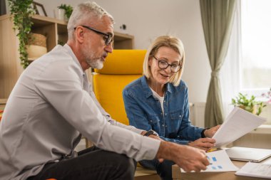 Olgun yaşlı çift karı koca birlikte çalışırlar. Evlerinde birlikte çalışırlar. Erkek ve kadın belgelere bakar ve konuşurlar.