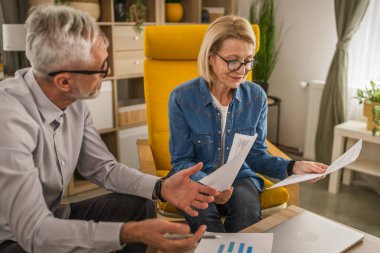Olgun yaşlı çift karı koca birlikte çalışırlar. Evlerinde birlikte çalışırlar. Erkek ve kadın belgelere bakar ve konuşurlar.