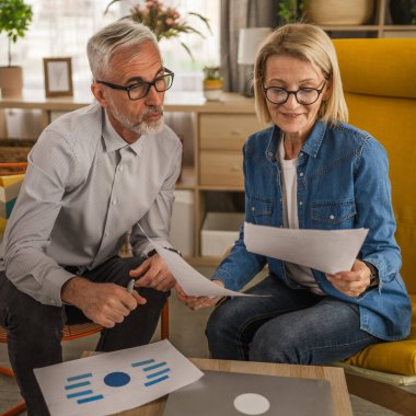 Olgun yaşlı çift karı koca birlikte çalışırlar. Evlerinde birlikte çalışırlar. Erkek ve kadın belgelere bakar ve konuşurlar.