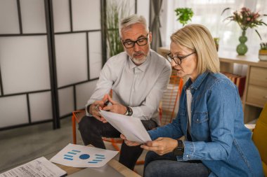 Olgun yaşlı çift karı koca birlikte çalışırlar. Evlerinde birlikte çalışırlar. Erkek ve kadın belgelere bakar ve konuşurlar.