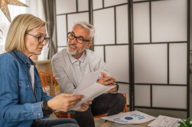 Olgun yaşlı çift karı koca birlikte çalışırlar. Evlerinde birlikte çalışırlar. Erkek ve kadın belgelere bakar ve konuşurlar.