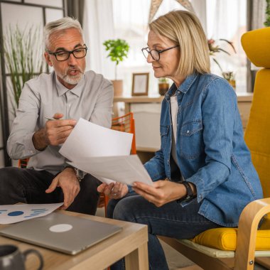 Olgun yaşlı çift karı koca birlikte çalışırlar. Evlerinde birlikte çalışırlar. Erkek ve kadın belgelere bakar ve konuşurlar.