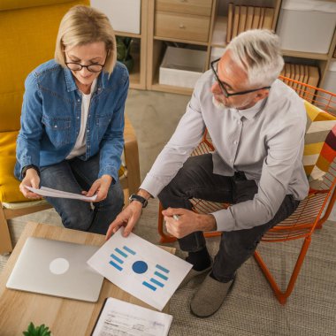 Olgun yaşlı çift karı koca birlikte çalışırlar. Evlerinde birlikte çalışırlar. Erkek ve kadın belgelere bakar ve konuşurlar.