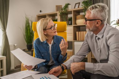 Olgun yaşlı çift karı koca birlikte çalışırlar. Evlerinde birlikte çalışırlar. Erkek ve kadın belgelere bakar ve konuşurlar.