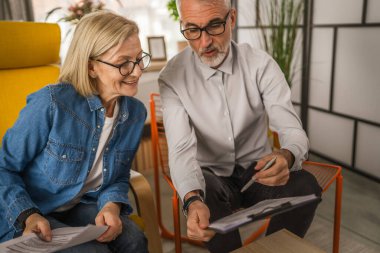 Olgun yaşlı çift karı koca birlikte çalışırlar. Evlerinde birlikte çalışırlar. Erkek ve kadın belgelere bakar ve konuşurlar.