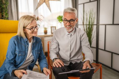 Olgun yaşlı çift karı koca birlikte çalışırlar. Evlerinde birlikte çalışırlar. Erkek ve kadın belgelere bakar ve konuşurlar.