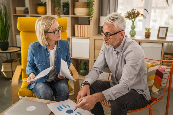 Olgun yaşlı çift karı koca birlikte çalışırlar. Evlerinde birlikte çalışırlar. Erkek ve kadın belgelere bakar ve konuşurlar.