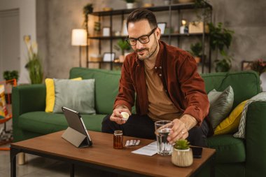 Yetişkin beyaz bir adam kanepede oturur ve evde ilaç içer.