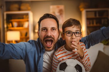 Evde futbol topu tezahüratı yapan baba ve oğulun portresi.