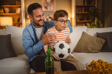 Yetişkin baba, futbol maçı izlerken elinde futbol topu tutan oğluna sarılıyor.