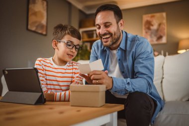Baba ve oğul evde tabletle birlikte hediye kutusundan kart okurlar.