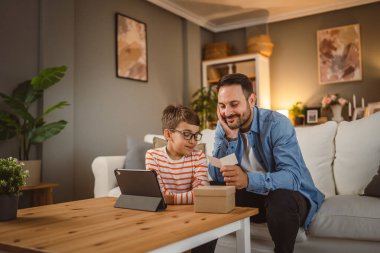 Baba ve oğul evde tabletle birlikte hediye kutusundan kart okurlar.