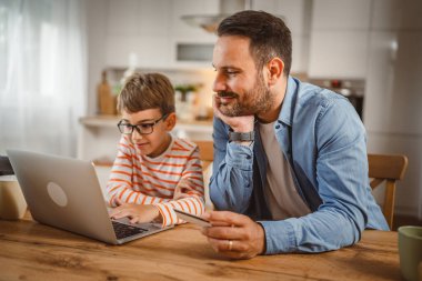 Çocuk mutfaktan dizüstü bilgisayar alırken, yetişkin beyaz bir babanın elinde kredi kartı var.