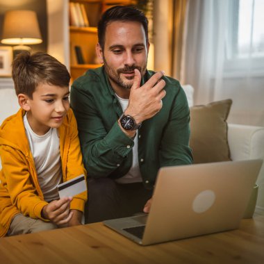 Beyaz çocuk, babasıyla kanepede oturur, kredi kartı alır, internetten alışveriş yapar, dizüstü bilgisayar alır.