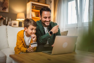 Beyaz çocuk, babasıyla kanepede oturur, kredi kartı alır, internetten alışveriş yapar, dizüstü bilgisayar alır.