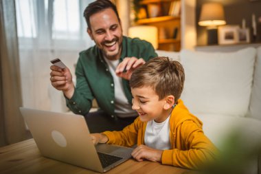 Kafkasyalı baba, oğluyla kanepede oturur, kredi kartı alır, bilgisayardan online alışveriş yapar.