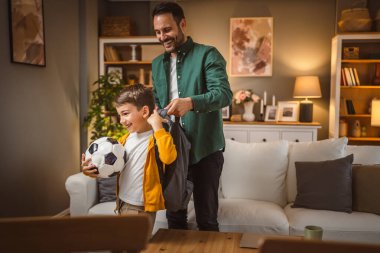 Baba, futbol topunu tutan oğluna okul çantasına koyması için yardım et.
