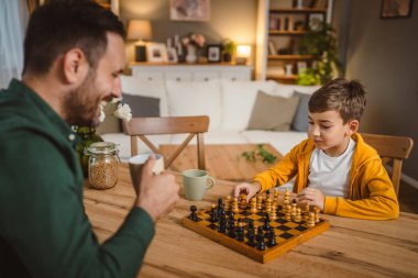 Bir bardak sıcak içecekle baba ve oğlu beyaz evde satranç oynuyor.
