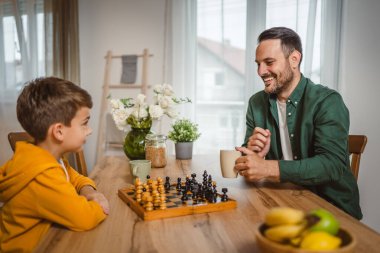 Bir bardak sıcak içecekle baba ve oğlu beyaz evde satranç oynuyor.