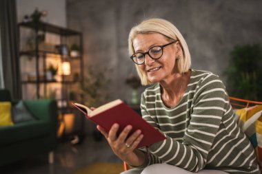 Olgun yaşlı kadın evde oturup kitap okuyor.