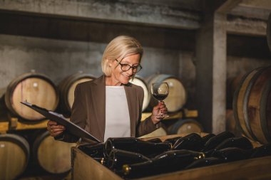 Olgun yaşlı şarap imalatçısı bodrumda duruyor. Elinde dosya ve bir kadeh şarap var.