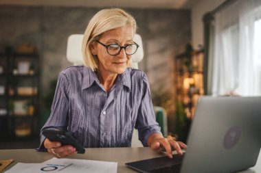 Olgun yaşlı kadın evde çalışıyor ve cep telefonu ve laptop kullanıyor.