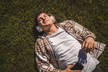 Kulaklıklı genç bir çocuk çimlere uzanıp cep telefonuyla müzik dinliyor.